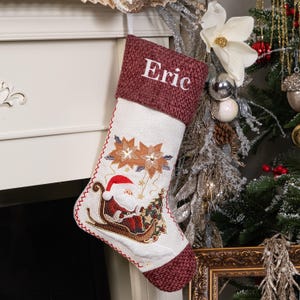 Puede incluir: Un calcet&iacute;n navide&ntilde;o con un pu&ntilde;o burdeos bordado con el nombre "Eric". El cuerpo blanco presenta bordados de flores de Pascua y Pap&aacute; Noel en un trineo con un &aacute;rbol de Navidad. El calcet&iacute;n est&aacute; decorado con costuras rojas.