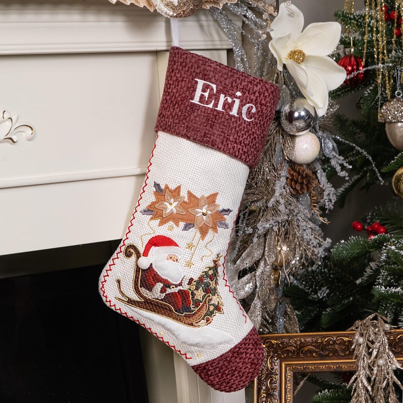 Puede incluir: Un calcet&iacute;n navide&ntilde;o con un pu&ntilde;o burdeos bordado con el nombre "Eric". El cuerpo blanco presenta bordados de flores de Pascua y Pap&aacute; Noel en un trineo con un &aacute;rbol de Navidad. El calcet&iacute;n est&aacute; decorado con costuras rojas.