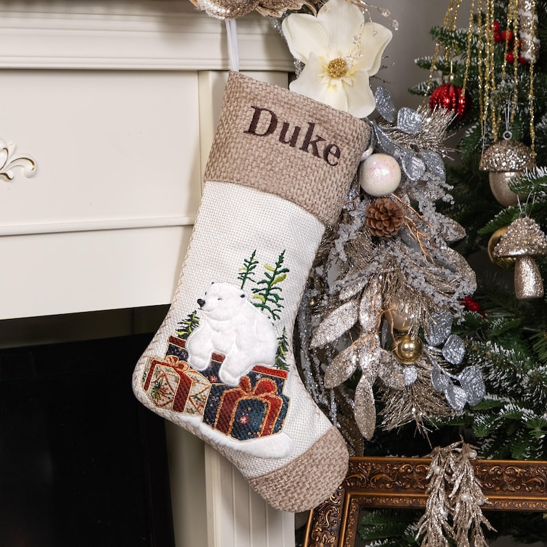 Puede incluir: Un calcet&iacute;n navide&ntilde;o con un dise&ntilde;o de oso polar. El calcet&iacute;n es blanco con un pu&ntilde;o marr&oacute;n y presenta un oso polar sentado encima de regalos envueltos. El nombre "Duke" est&aacute; bordado en el pu&ntilde;o. Hay &aacute;rboles verdes bordados en el calcet&iacute;n.
