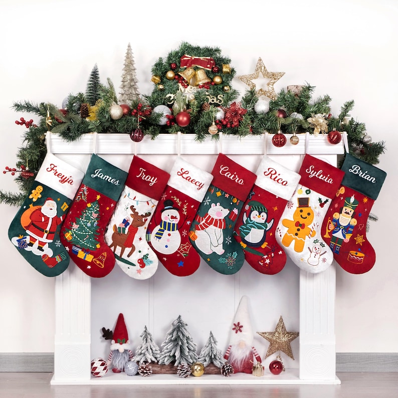 May include: Eight personalized Christmas stockings with embroidered names and festive designs. The stockings are hung on a white fireplace mantel with a green garland and red ornaments. The stockings feature designs such as Santa Claus, a snowman, a penguin, a gingerbread man, and a nutcracker. The names on the stockings are Frejya, James, Troy, Lacey, Cairo, Rico, Sylvia, and Brian.