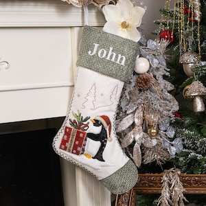 Puede incluir: Un calcet&iacute;n navide&ntilde;o con un dise&ntilde;o de ping&uuml;ino. El calcet&iacute;n es blanco con la parte superior y el ribete verdes. El ping&uuml;ino lleva un gorro de Pap&aacute; Noel y sostiene un regalo. El nombre "John" est&aacute; bordado.