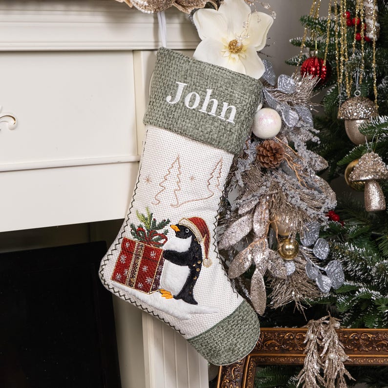 Puede incluir: Un calcet&iacute;n navide&ntilde;o con un dise&ntilde;o de ping&uuml;ino. El calcet&iacute;n es blanco con la parte superior y el ribete verdes. El ping&uuml;ino lleva un gorro de Pap&aacute; Noel y sostiene un regalo. El nombre "John" est&aacute; bordado.