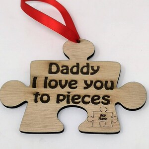 Puede incluir: Adorno de madera en forma de pieza de rompecabezas con el texto "Daddy I love you to pieces" y una pieza de rompecabezas más pequeña con el texto "Any Name". El adorno está sujeto a una cinta roja.