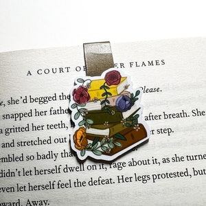 Peut inclure: Une pile de livres avec des accents floraux, un marque-page et le texte "A Court of Silver Flames" et "Please." Le marque-page est un clip en métal doré.