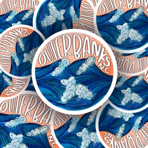 May include: Close-up of multiple stickers with a watercolor design of blue waves and white clouds. The stickers say "Outer Banks NC" in white lettering on a peach background.