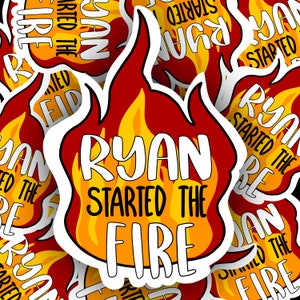 Puede incluir: Una pegatina de llama roja y amarilla con el texto "Ryan Started the Fire" en blanco. La pegatina es un diseño popular para los fanáticos de la canción "Started the Fire" de Billy Joel.