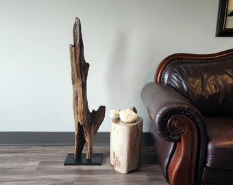 May include: A tall, brown driftwood sculpture stands on a black base next to a wooden stool with a white, organic-shaped object on top. A brown leather sofa with a wooden frame is in the background.