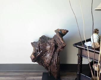 May include: A large, dark brown, wooden sculpture of a tree trunk with a natural, rough texture. The sculpture is mounted on a black base and stands on a wood floor.