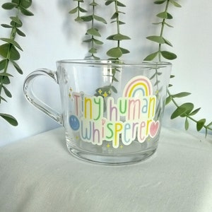 May include: A clear glass mug with a rainbow and the text "Tiny human whisperer" in colourful letters. The mug has a smiley face and a heart on either side of the text.