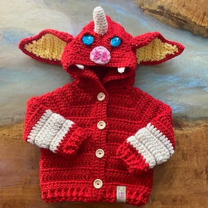 May include: Red crocheted baby cardigan with a monster hood featuring a white horn, pink nose, and blue eyes. The cardigan has white and red striped cuffs and buttons down the front.