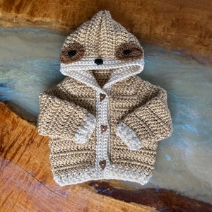 May include: A crocheted baby cardigan with a sloth face on the hood. The cardigan is beige and white with brown felt eyes and a brown felt nose. The cardigan has a button closure with brown heart-shaped buttons.