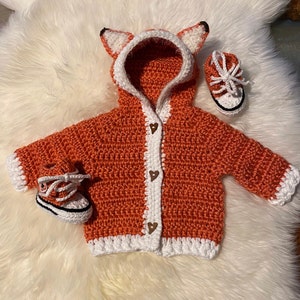 K&ouml;nnte beinhalten: Ein geh&auml;kelter orangefarbener Fuchs-Kapuzenpullover mit wei&szlig;em Besatz und braunen Herz-Kn&ouml;pfen. Der Pullover wird mit zwei geh&auml;kelten orangefarbenen und wei&szlig;en Schuhen geliefert.