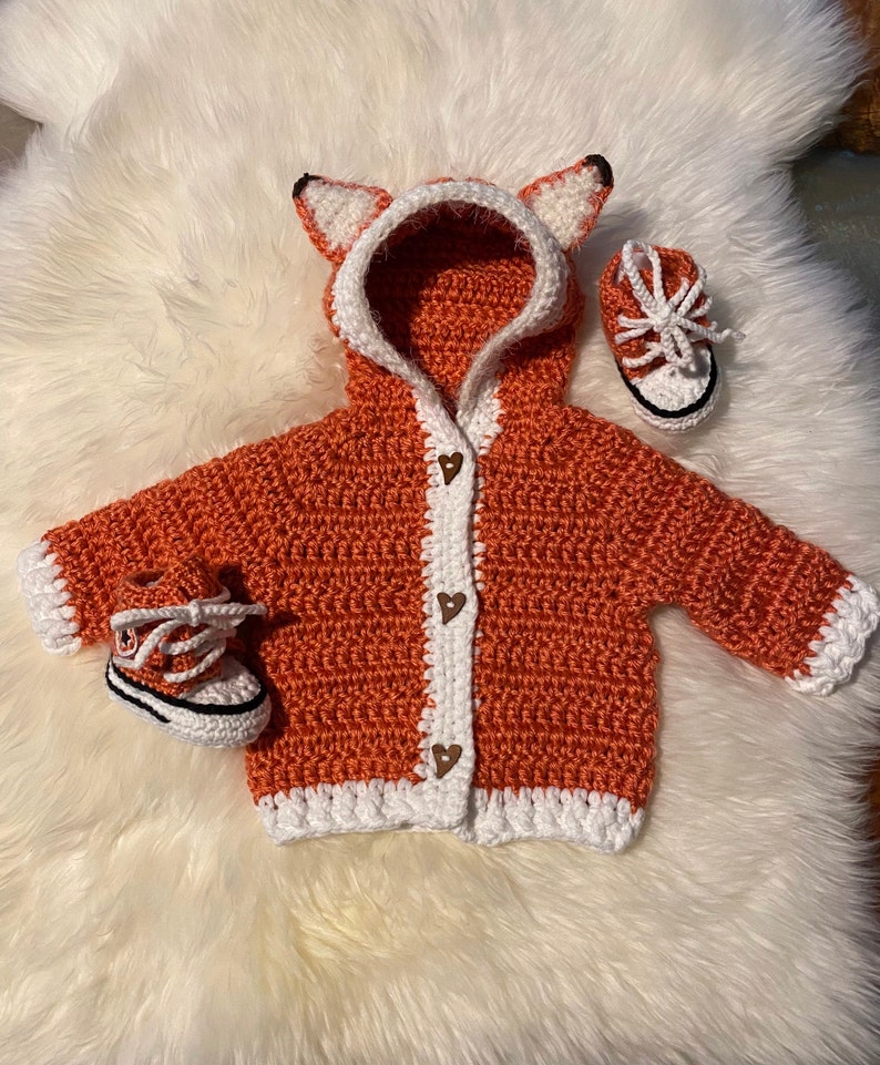 K&ouml;nnte beinhalten: Ein geh&auml;kelter orangefarbener Fuchs-Kapuzenpullover mit wei&szlig;em Besatz und braunen Herz-Kn&ouml;pfen. Der Pullover wird mit zwei geh&auml;kelten orangefarbenen und wei&szlig;en Schuhen geliefert.