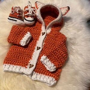 K&ouml;nnte beinhalten: Ein geh&auml;kelter orangefarbener Fuchs-Cardigan mit wei&szlig;em Besatz und Holz-Herz-Kn&ouml;pfen. Der Cardigan hat eine Kapuze mit Fuchs-Ohren. Ein Paar orangefarbene und wei&szlig;e geh&auml;kelte Babyschuhe sind enthalten.