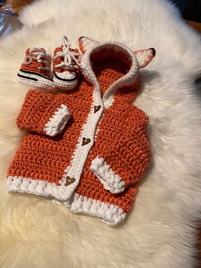 K&ouml;nnte beinhalten: Ein geh&auml;kelter orangefarbener Fuchs-Cardigan mit wei&szlig;em Besatz und Holz-Herz-Kn&ouml;pfen. Der Cardigan hat eine Kapuze mit Fuchs-Ohren. Ein Paar orangefarbene und wei&szlig;e geh&auml;kelte Babyschuhe sind enthalten.