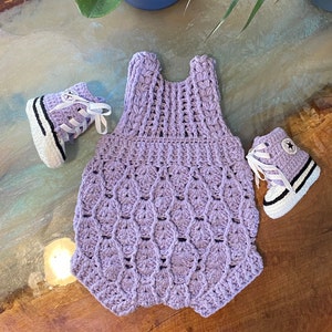 May include: A lavender crocheted baby onesie with a scalloped design and matching crocheted baby shoes. The shoes have white laces and black trim.