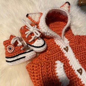 K&ouml;nnte beinhalten: Ein geh&auml;keltes Fuchs-Kost&uuml;m f&uuml;r ein Baby, bestehend aus einer Kapuzenjacke mit wei&szlig;em Besatz und zwei geh&auml;kelten Converse-Schuhen.