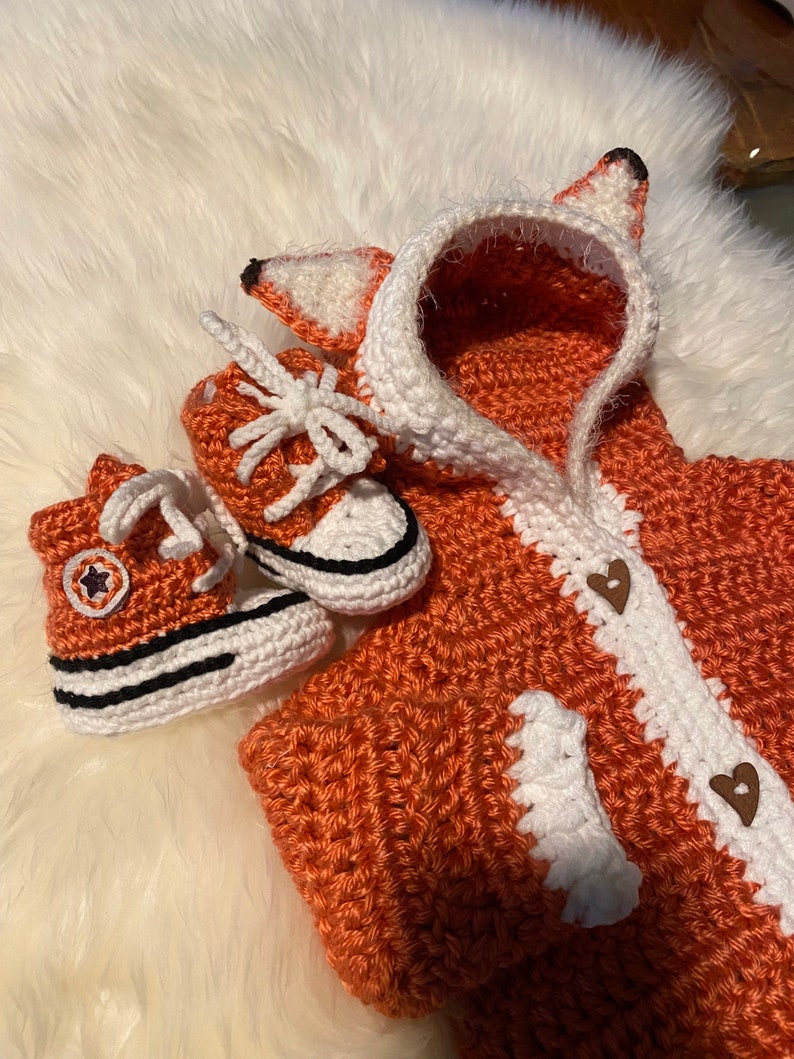 K&ouml;nnte beinhalten: Ein geh&auml;keltes Fuchs-Kost&uuml;m f&uuml;r ein Baby, bestehend aus einer Kapuzenjacke mit wei&szlig;em Besatz und zwei geh&auml;kelten Converse-Schuhen.