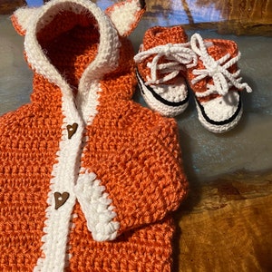 K&ouml;nnte beinhalten: Ein geh&auml;kelter orangefarbener Fuchs-Kapuzenpullover mit wei&szlig;em Besatz und Holzknopf-Herzen. Der Pullover wird mit einem Paar geh&auml;kelter orangefarbener und wei&szlig;er Babyschuhe geliefert.