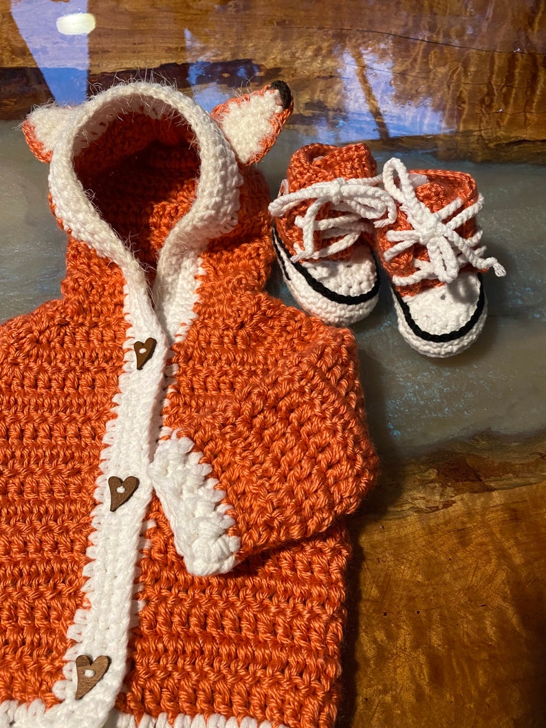 K&ouml;nnte beinhalten: Ein geh&auml;kelter orangefarbener Fuchs-Kapuzenpullover mit wei&szlig;em Besatz und Holzknopf-Herzen. Der Pullover wird mit einem Paar geh&auml;kelter orangefarbener und wei&szlig;er Babyschuhe geliefert.