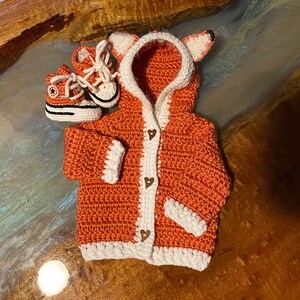K&ouml;nnte beinhalten: Ein geh&auml;kelter orangefarbener Fuchs-Cardigan mit wei&szlig;em Besatz und herzf&ouml;rmigen Kn&ouml;pfen. Der Cardigan hat eine Kapuze mit Fuchs-Ohren. Zwei kleine geh&auml;kelte orangefarbene und wei&szlig;e Schuhe sind im Lieferumfang enthalten.