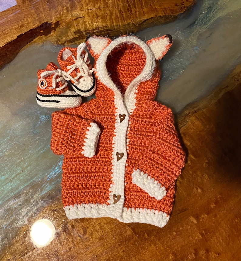 K&ouml;nnte beinhalten: Ein geh&auml;kelter orangefarbener Fuchs-Cardigan mit wei&szlig;em Besatz und herzf&ouml;rmigen Kn&ouml;pfen. Der Cardigan hat eine Kapuze mit Fuchs-Ohren. Zwei kleine geh&auml;kelte orangefarbene und wei&szlig;e Schuhe sind im Lieferumfang enthalten.