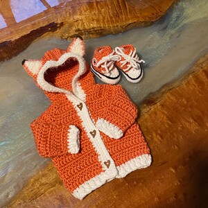 K&ouml;nnte beinhalten: Ein geh&auml;kelter Fuchs-Kapuzenpullover in Orange mit wei&szlig;em Besatz und braunen Herz-Kn&ouml;pfen. Der Pullover liegt auf einer Holzoberfl&auml;che mit einem Paar geh&auml;kelter Babyschuhe.
