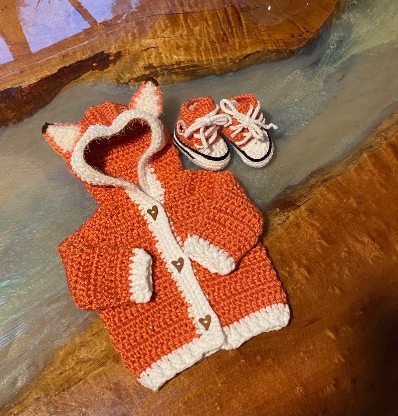 K&ouml;nnte beinhalten: Ein geh&auml;kelter Fuchs-Kapuzenpullover in Orange mit wei&szlig;em Besatz und braunen Herz-Kn&ouml;pfen. Der Pullover liegt auf einer Holzoberfl&auml;che mit einem Paar geh&auml;kelter Babyschuhe.