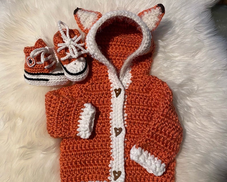 K&ouml;nnte beinhalten: Ein geh&auml;kelter orangefarbener Fuchs-Kapuzenpullover mit wei&szlig;en Verzierungen und einem Knopfverschluss. Der Pullover wird mit zwei geh&auml;kelten orangefarbenen und wei&szlig;en Babyschuhen kombiniert.