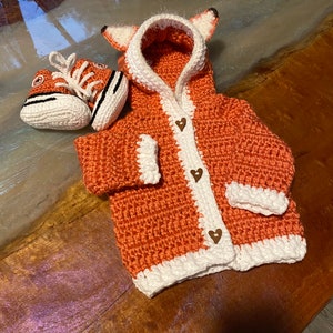 K&ouml;nnte beinhalten: Ein geh&auml;kelter orangefarbener und wei&szlig;er Fuchs-Kapuzenpullover mit Holz-Herz-Kn&ouml;pfen. Der Pullover wird mit einem geh&auml;kelten orangefarbenen und wei&szlig;en Paar Babyschuhe kombiniert.