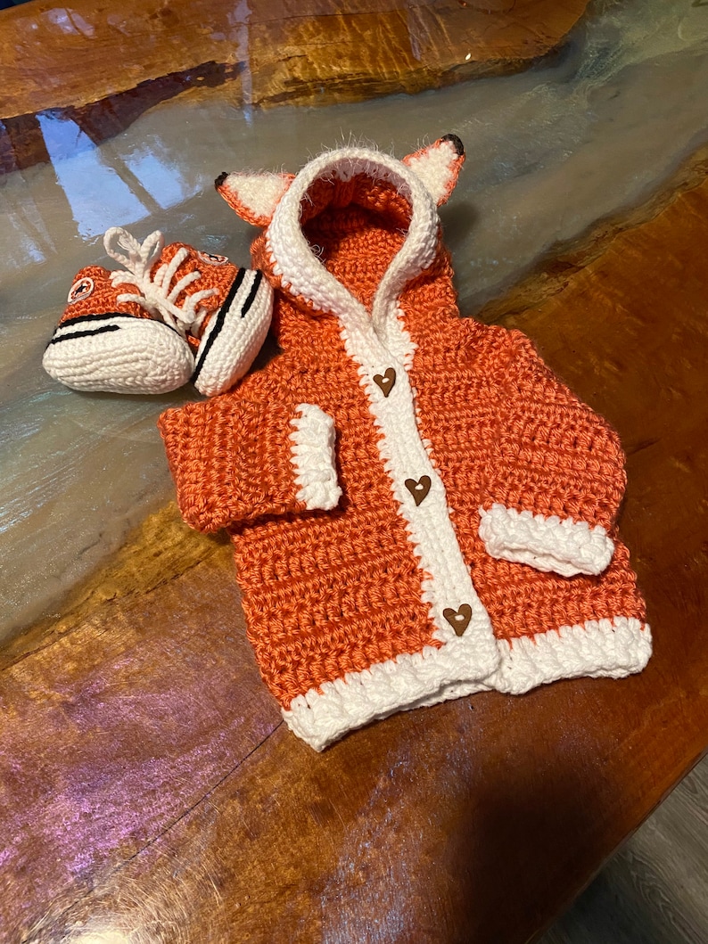 K&ouml;nnte beinhalten: Ein geh&auml;kelter orangefarbener und wei&szlig;er Fuchs-Kapuzenpullover mit Holz-Herz-Kn&ouml;pfen. Der Pullover wird mit einem geh&auml;kelten orangefarbenen und wei&szlig;en Paar Babyschuhe kombiniert.