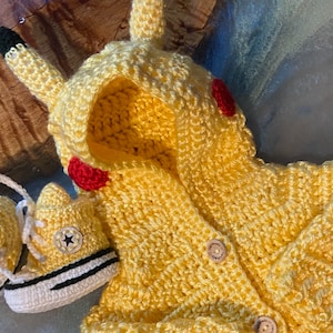 May include: Yellow crocheted Pikachu baby outfit with a hood and a button closure. The outfit is paired with two yellow crocheted baby shoes with black trim.