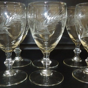 Può includere: Set di sei bicchieri da vino in vetro trasparente con un motivo inciso di spighe di grano. I bicchieri hanno una coppa rotonda e un gambo alto e sottile.