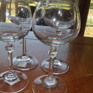 May include: Four clear glass wine glasses with a round bowl and a long stem. The glasses are sitting on a wooden table.