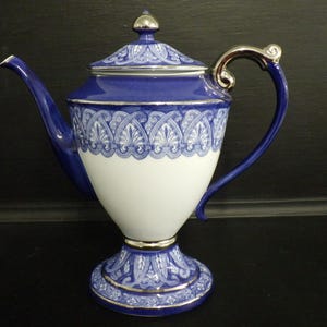 Op de afbeelding: Een blauw-witte porseleinen koffiepot met een zilverkleurige handgreep en dekselknop. De pot heeft een decoratief patroon rond de voet, de body en het deksel. De handgreep en tuit zijn blauw en de body is wit.