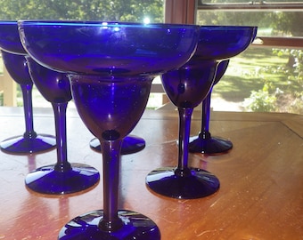 May include: Five cobalt blue margarita glasses on a wooden table. The glasses are empty and have a wide rim.