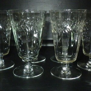 May include: A set of six clear stemmed glasses. Each glass has a textured, vertical pattern on the bowl and a circular base. The glasses are arranged on a dark surface, highlighting their elegant design.