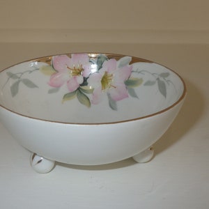 Puede incluir: Un tazón de porcelana blanca con un borde dorado y un diseño floral. El tazón tiene tres pequeñas patas.