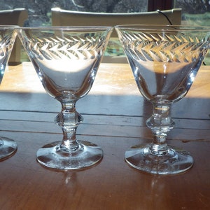 May include: Four clear glass wine glasses with a decorative etched design of a repeating pattern of leaves and arrows. The glasses are on a wooden table.