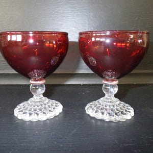 Pode incluir: Dois copos de vinho tintos vermelho rubi. As taças são de um vermelho profundo, e as hastes são transparentes com uma pequena base redonda. A base de cada copo é transparente e tem um padrão texturizado em forma de bolhas. Os copos estão sobre um fundo escuro.