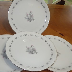 May include: Four white ceramic plates with a floral design. Each plate has a central floral motif and a border of small, stylized leaves. The plates have a gold rim and are arranged on a wooden surface.