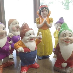 以下が含まれることがあります： 白雪姫と七人の小人を描いた陶器製のフィギュアのコレクション。フィギュアは赤、黄色、緑、紫などの鮮やかな色で塗装されています。フィギュアは木製のテーブルの上に配置されています。