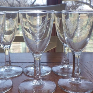 Op de afbeelding: Zes heldere glazen wijnglazen met gegraveerde ontwerpen aan de zijkanten. De glazen zijn in een halve cirkel op een houten tafel gerangschikt.