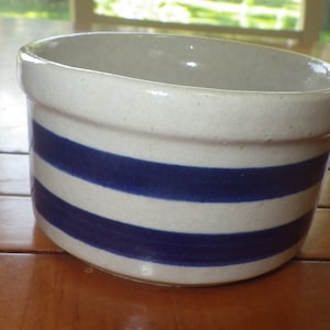 May include: A small white ceramic crock with blue stripes. The crock is round and has a wide opening.