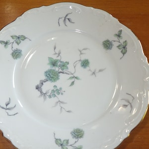 May include: A white porcelain plate with a gold rim and a floral design in shades of green. The plate features a delicate floral pattern with leaves and flowers.