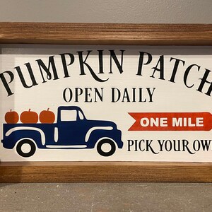 May include: A wooden sign with a white background and black text that reads "Pumpkin Patch Open Daily One Mile Pick Your Own". A blue truck with a bed full of orange pumpkins is pictured in the center of the sign.