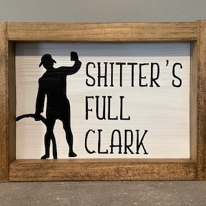 May include: A wooden sign with a white background and black text that reads "Shitter's Full Clark". A silhouette of a person holding a beer mug is to the left of the text.