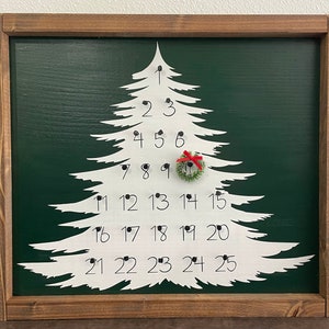 May include: A white wooden Christmas tree cutout with numbers 1 through 25 on it. The tree is mounted on a green background and framed in brown wood.