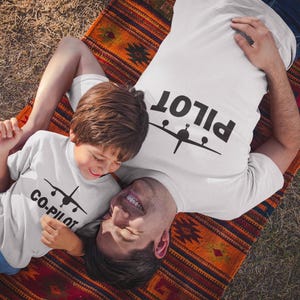 May include: A father and son wearing matching white t-shirts with a plane graphic. The father's shirt says "PILOT" and the son's shirt says "CO-PILOT". They are lying on a colourful rug.