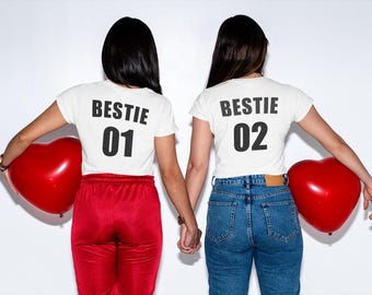 Puede incluir: Dos personas con camisetas blancas con el texto "Bestie 01" y "Bestie 02" en la espalda. Están sosteniendo globos rojos en forma de corazón.